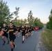 A large group of U.S. Army personnel are conducting a formation run along a paved road flanked by trees. All participants are wearing matching black PT (Physical Training) uniforms with 'ARMY' printed in gold/yellow lettering. The photo appears to be taken during early morning or evening hours based on the soft, low-angle lighting.