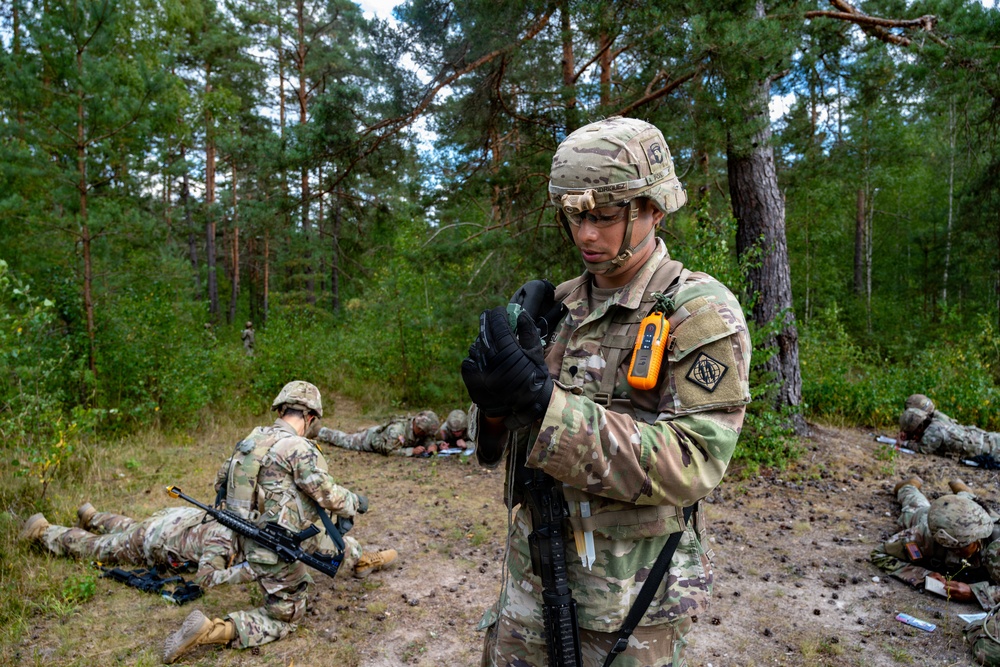 A U.S. Army soldier in MultiCam/OCP camouflage uniform stands in a forested training area, putting on black tactical gloves while carrying a rifle. Several other soldiers are visible in the background, some lying on the ground in what appears to be a field training exercise. The setting appears to be a pine forest, consistent with Central or Eastern European terrain.