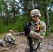 A U.S. Army soldier in MultiCam/OCP camouflage uniform stands in a forested training area, putting on black tactical gloves while carrying a rifle. Several other soldiers are visible in the background, some lying on the ground in what appears to be a field training exercise. The setting appears to be a pine forest, consistent with Central or Eastern European terrain.