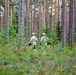 Two military personnel in camouflage uniforms and helmets are patrolling through a dense coniferous forest. They are moving away from the camera through undergrowth and low vegetation. The setting appears to be a northern European pine forest based on the tall, slender tree trunks and ground cover.