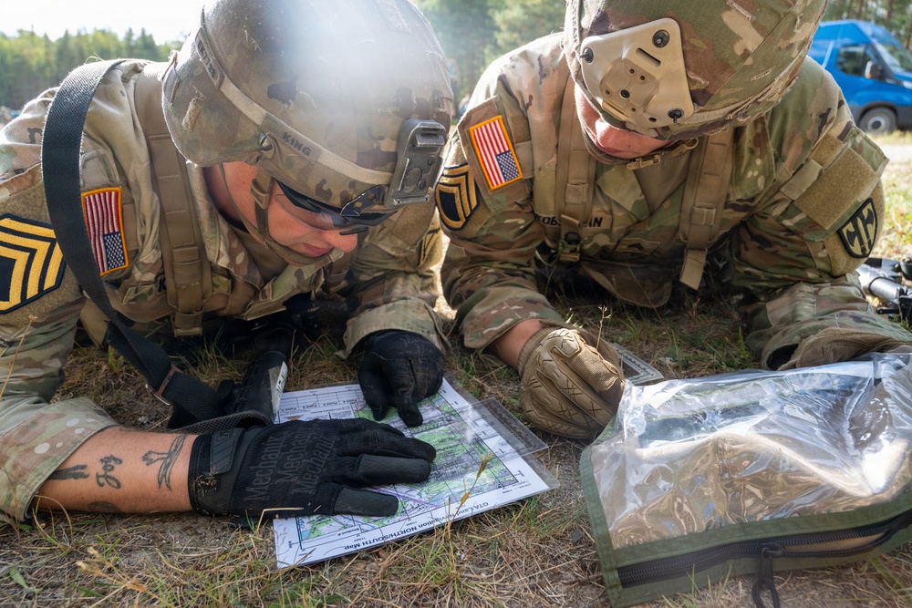 Two U.S. Army soldiers in full combat gear are lying prone on the ground studying a topographic map. Both wear OCP (Operational Camouflage Pattern) uniforms with American flag patches and Staff Sergeant rank insignia. They appear to be conducting land navigation or mission planning in an outdoor field environment.