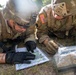 Two U.S. Army soldiers in full combat gear are lying prone on the ground studying a topographic map. Both wear OCP (Operational Camouflage Pattern) uniforms with American flag patches and Staff Sergeant rank insignia. They appear to be conducting land navigation or mission planning in an outdoor field environment.