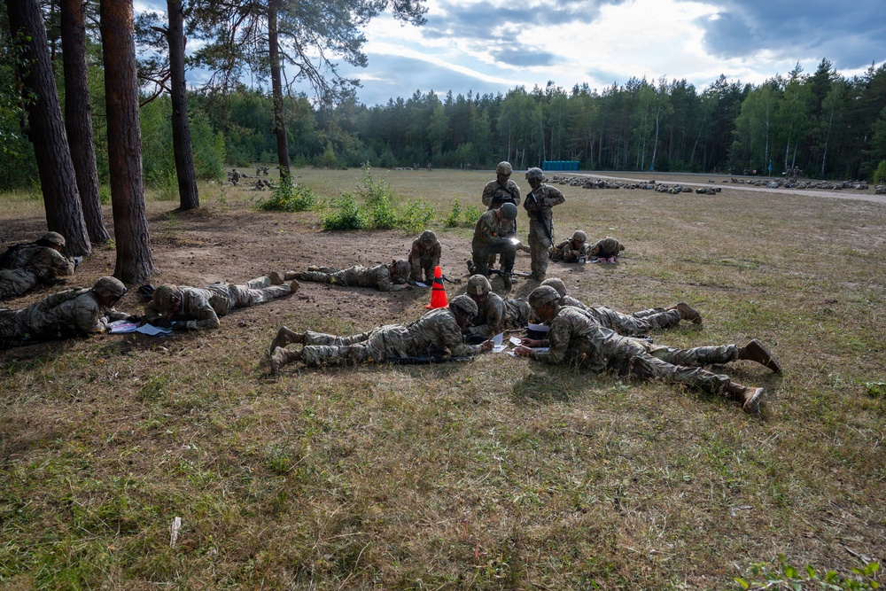 A group of military personnel in camouflage uniforms are conducting a field training exercise on an open grassy area bordered by pine and birch trees. Several soldiers are in prone positions on the ground appearing to review documents or maps, while others are standing or kneeling in a supervisory or instructional role. The setting appears to be a military training range, evidenced by the open field, a portable toilet in the background, and what appear to be additional soldiers or targets visible in the distance.