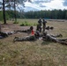 A group of military personnel in camouflage uniforms are conducting a field training exercise on an open grassy area bordered by pine and birch trees. Several soldiers are in prone positions on the ground appearing to review documents or maps, while others are standing or kneeling in a supervisory or instructional role. The setting appears to be a military training range, evidenced by the open field, a portable toilet in the background, and what appear to be additional soldiers or targets visible in the distance.