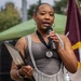 A woman is speaking into a microphone at an outdoor event, holding what appears to be an official certificate or award folder. She is wearing a medal on a lanyard around her neck, suggesting she is being honored or is presenting at a formal ceremony. The setting includes a green canopy tent and a maroon and gold flag in the background.