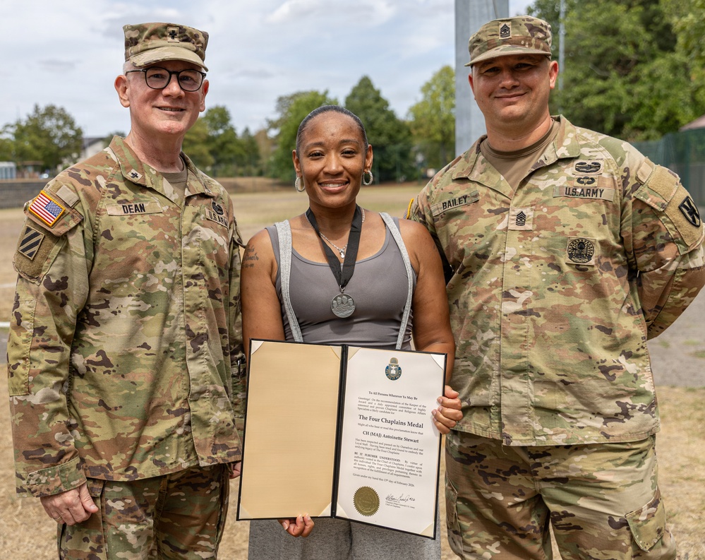 Three individuals pose for a photo outdoors, with a civilian woman in the center holding an open award certificate. The document visible is 'The Four Chaplains Medal' awarded to 'CH (MAJ) Antoinette Stewart.' She is flanked by two U.S. Army soldiers in OCP (Operational Camouflage Pattern) uniforms, identified by name tapes as 'DEAN' on the left and 'BAILEY' on the right.
