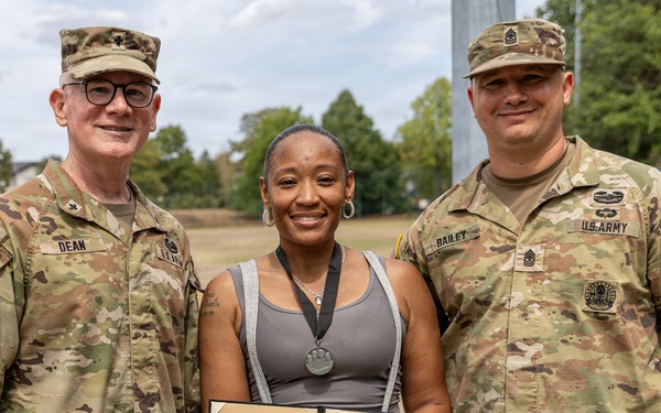 Three individuals pose for a photo outdoors, with a civilian woman in the center holding an open award certificate. The document visible is 'The Four Chaplains Medal' awarded to 'CH (MAJ) Antoinette Stewart.' She is flanked by two U.S. Army soldiers in OCP (Operational Camouflage Pattern) uniforms, identified by name tapes as 'DEAN' on the left and 'BAILEY' on the right.