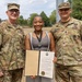 Three individuals pose for a photo outdoors, with a civilian woman in the center holding an open award certificate. The document visible is 'The Four Chaplains Medal' awarded to 'CH (MAJ) Antoinette Stewart.' She is flanked by two U.S. Army soldiers in OCP (Operational Camouflage Pattern) uniforms, identified by name tapes as 'DEAN' on the left and 'BAILEY' on the right.