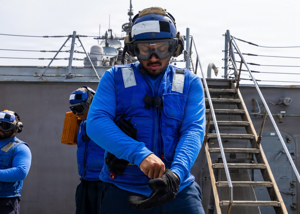 A naval sailor dressed in blue flight deck gear is on the deck of a military vessel, appearing to don or adjust gloves and equipment. At least two other similarly dressed crew members are visible in the background. The setting is clearly a warship deck with metal staircases, rigging, and superstructure visible.