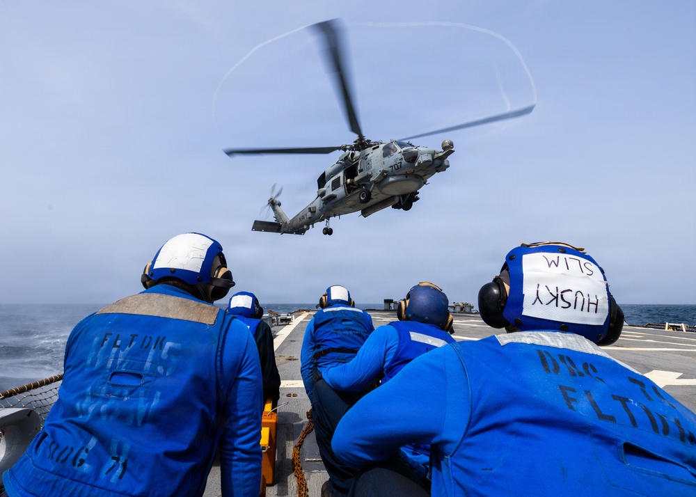 Naval personnel in blue flight deck uniforms observe a military helicopter hovering over a ship's flight deck at sea. The helicopter, numbered '707', appears to be conducting a landing or hover operation above the deck. Crew members are wearing hearing protection and have role/name markings on their helmets and jackets.