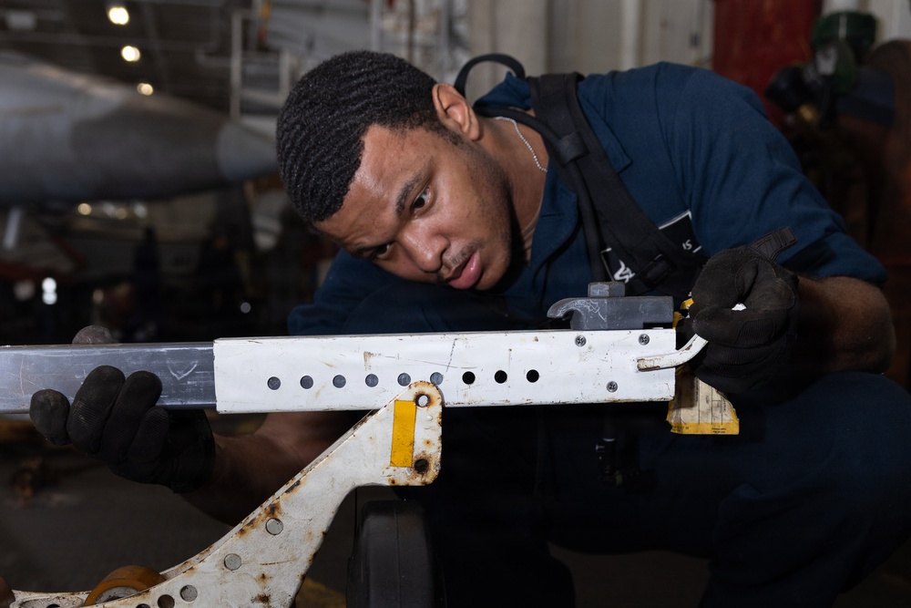 A young male technician wearing a dark blue uniform and black gloves is closely inspecting or working on a white metal component, likely an aircraft part. He is leaning in to examine the piece carefully, which is mounted on a worn metal stand with visible rust. The setting appears to be an aircraft hangar or maintenance bay, with aircraft visible in the background.