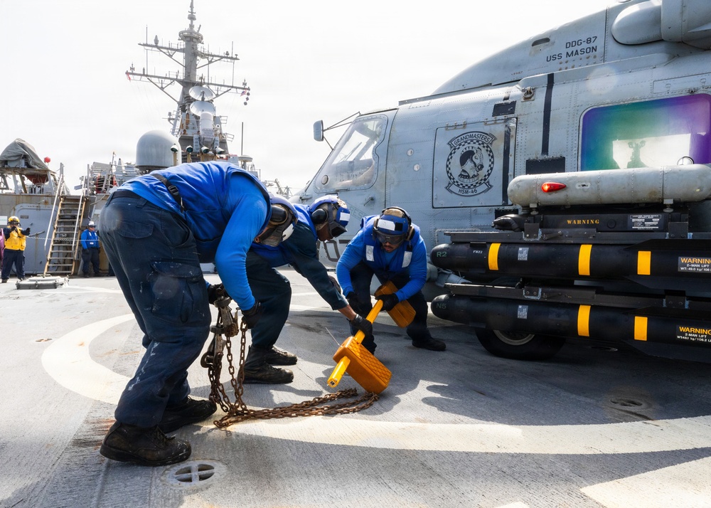 Three Navy sailors in blue coveralls are working on the flight deck of USS Mason (DDG-87), securing or unsecuring a helicopter with chains and wheel chocks. The helicopter bears the markings of HSM-46 'Grandmasters' squadron. The scene takes place on the ship's helicopter landing pad, with the destroyer's superstructure visible in the background.