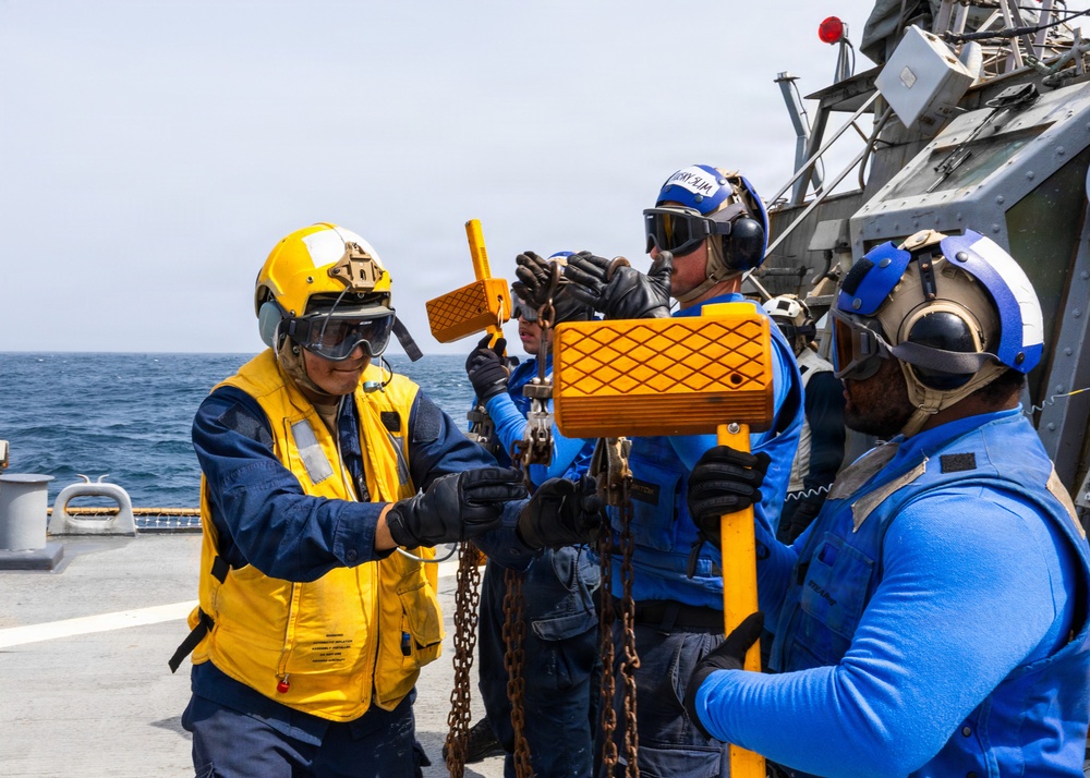 Naval personnel are working on the deck of a military vessel at sea, handling chains and orange-colored equipment that appears to be related to deck operations or replenishment-at-sea procedures. The crew members are wearing standard naval flight deck safety gear including helmets, goggles, hearing protection, and color-coded vests and shirts indicating their roles. The scene takes place in clear daylight conditions with open ocean visible in the background.