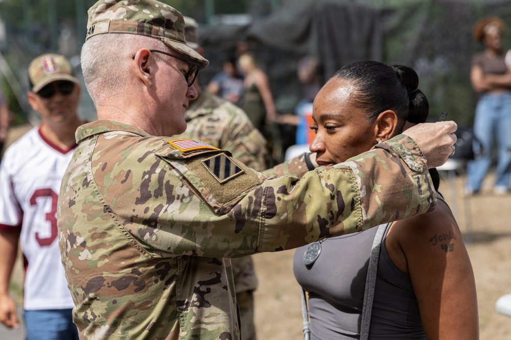 A U.S. Army soldier in OCP (Operational Camouflage Pattern) uniform is pinning a medal or insignia onto a civilian woman in a gray top at what appears to be an outdoor military event or ceremony. The soldier wears a U.S. flag patch and a 3rd Infantry Division patch on his sleeve. The setting appears to be a public or community event with bystanders visible in the background.