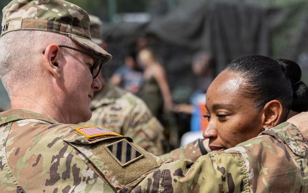 A U.S. Army soldier in OCP (Operational Camouflage Pattern) uniform is pinning a medal or insignia onto a civilian woman in a gray top at what appears to be an outdoor military event or ceremony. The soldier wears a U.S. flag patch and a 3rd Infantry Division patch on his sleeve. The setting appears to be a public or community event with bystanders visible in the background.