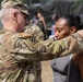 A U.S. Army soldier in OCP (Operational Camouflage Pattern) uniform is pinning a medal or insignia onto a civilian woman in a gray top at what appears to be an outdoor military event or ceremony. The soldier wears a U.S. flag patch and a 3rd Infantry Division patch on his sleeve. The setting appears to be a public or community event with bystanders visible in the background.