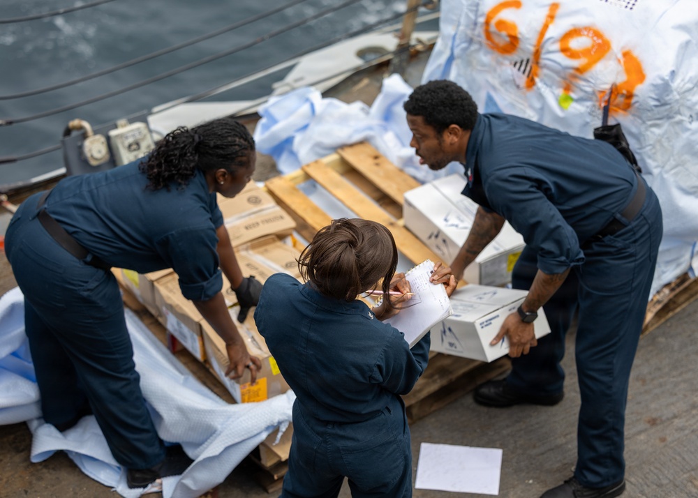 Three individuals wearing matching dark navy blue uniforms are gathered around a wooden pallet on what appears to be a ship's deck. They are inspecting and documenting boxes and cargo items, with one person writing on a clipboard or notepad. A large white tarp or covering with orange markings is visible in the background.