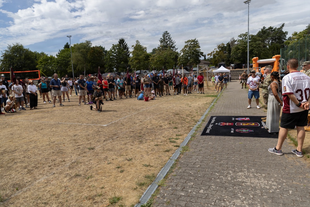 A large outdoor gathering of people at what appears to be a military community event or sports day, held on a dry grass field. A crowd of approximately 50-100 people lines the perimeter of the field, with some individuals in military camouflage uniforms and others in civilian clothing including sports jerseys numbered '30'. A branded mat/banner with military insignia is visible on a paved pathway in the foreground.
