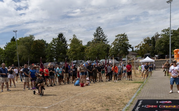 A large outdoor gathering of people at what appears to be a military community event or sports day, held on a dry grass field. A crowd of approximately 50-100 people lines the perimeter of the field, with some individuals in military camouflage uniforms and others in civilian clothing including sports jerseys numbered '30'. A branded mat/banner with military insignia is visible on a paved pathway in the foreground.