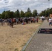 A large outdoor gathering of people at what appears to be a military community event or sports day, held on a dry grass field. A crowd of approximately 50-100 people lines the perimeter of the field, with some individuals in military camouflage uniforms and others in civilian clothing including sports jerseys numbered '30'. A branded mat/banner with military insignia is visible on a paved pathway in the foreground.