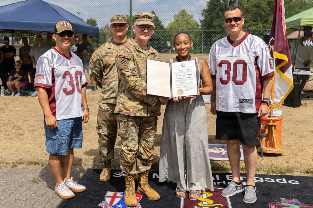 A military award ceremony is taking place outdoors, where two soldiers in Army Combat Uniforms are presenting what appears to be a 'Four Chaplains Medal' certificate to a civilian woman. The group of five individuals includes two men wearing matching white and maroon number 30 football jerseys labeled 'Victory Rider.' The event appears to be associated with the 3rd Infantry Brigade, as indicated by the mat visible on the ground.