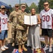 A military award ceremony is taking place outdoors, where two soldiers in Army Combat Uniforms are presenting what appears to be a 'Four Chaplains Medal' certificate to a civilian woman. The group of five individuals includes two men wearing matching white and maroon number 30 football jerseys labeled 'Victory Rider.' The event appears to be associated with the 3rd Infantry Brigade, as indicated by the mat visible on the ground.