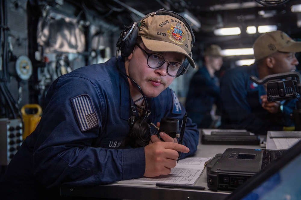 A U.S. Navy sailor is stationed at a workstation aboard the USS San Diego (LPD-22), wearing a ship's ball cap and communication headset while holding a handset and reviewing documents. The setting appears to be the ship's bridge or combat information center. At least one other crew member is visible in the background performing similar duties.