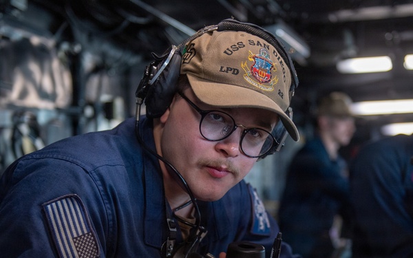 A U.S. Navy sailor is stationed at a workstation aboard the USS San Diego (LPD-22), wearing a ship's ball cap and communication headset while holding a handset and reviewing documents. The setting appears to be the ship's bridge or combat information center. At least one other crew member is visible in the background performing similar duties.