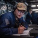 A U.S. Navy sailor is stationed at a workstation aboard the USS San Diego (LPD-22), wearing a ship's ball cap and communication headset while holding a handset and reviewing documents. The setting appears to be the ship's bridge or combat information center. At least one other crew member is visible in the background performing similar duties.