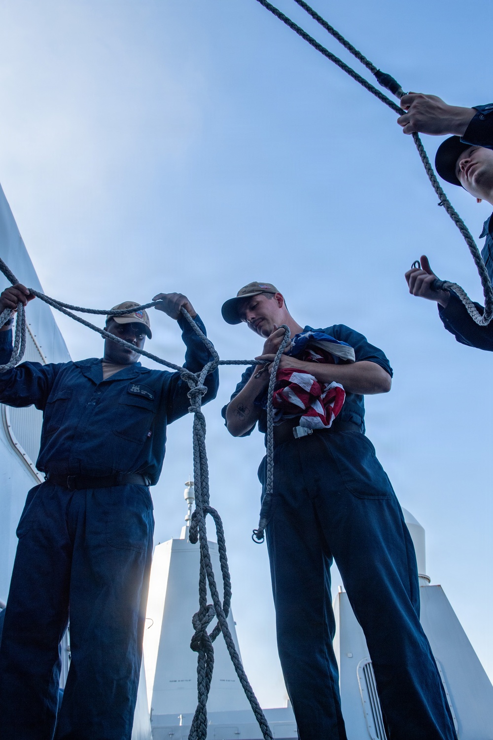 Three individuals in navy blue coveralls are working with ropes and halyards on the deck of a naval vessel. One person is holding a folded American flag while managing a line, suggesting a flag-raising or lowering ceremony. The low-angle shot looking upward emphasizes the rigging and open sky above.