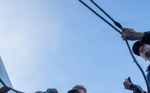 Three individuals in navy blue coveralls are working with ropes and halyards on the deck of a naval vessel. One person is holding a folded American flag while managing a line, suggesting a flag-raising or lowering ceremony. The low-angle shot looking upward emphasizes the rigging and open sky above.