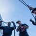 Three individuals in navy blue coveralls are working with ropes and halyards on the deck of a naval vessel. One person is holding a folded American flag while managing a line, suggesting a flag-raising or lowering ceremony. The low-angle shot looking upward emphasizes the rigging and open sky above.