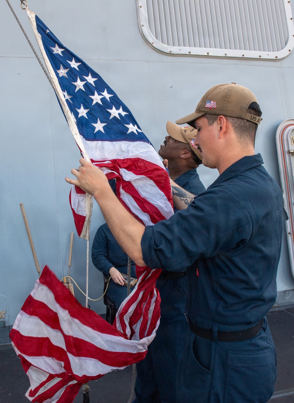 Two or three U.S. Navy sailors in Navy Working Uniform (NWU) are handling an American flag on the deck of a naval vessel. The foreground sailor wears a cap with 'LPD 22' visible, indicating assignment to USS San Diego (LPD-22). The sailors appear to be either raising or lowering the ensign during a colors ceremony.