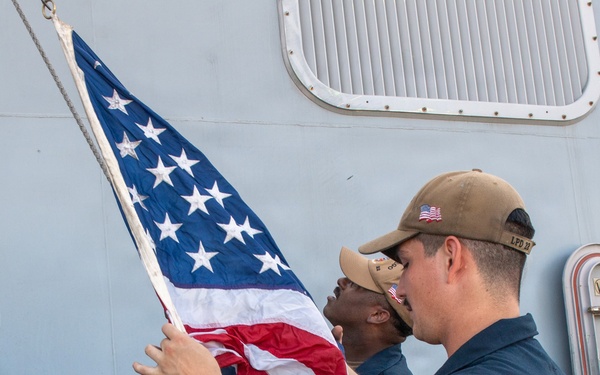 Two or three U.S. Navy sailors in Navy Working Uniform (NWU) are handling an American flag on the deck of a naval vessel. The foreground sailor wears a cap with 'LPD 22' visible, indicating assignment to USS San Diego (LPD-22). The sailors appear to be either raising or lowering the ensign during a colors ceremony.
