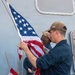 Two or three U.S. Navy sailors in Navy Working Uniform (NWU) are handling an American flag on the deck of a naval vessel. The foreground sailor wears a cap with 'LPD 22' visible, indicating assignment to USS San Diego (LPD-22). The sailors appear to be either raising or lowering the ensign during a colors ceremony.