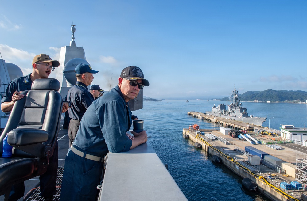 Military personnel in navy blue uniforms are observed on the bridge wing of a naval vessel, overlooking a harbor with a pier and a warship docked alongside. The setting appears to be a naval base with mountainous terrain visible in the background, suggesting a location in East Asia, possibly Japan. The scene depicts routine port operations or arrival/departure procedures.
