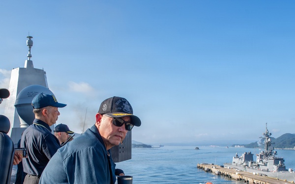 Military personnel in navy blue uniforms are observed on the bridge wing of a naval vessel, overlooking a harbor with a pier and a warship docked alongside. The setting appears to be a naval base with mountainous terrain visible in the background, suggesting a location in East Asia, possibly Japan. The scene depicts routine port operations or arrival/departure procedures.