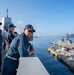 Military personnel in navy blue uniforms are observed on the bridge wing of a naval vessel, overlooking a harbor with a pier and a warship docked alongside. The setting appears to be a naval base with mountainous terrain visible in the background, suggesting a location in East Asia, possibly Japan. The scene depicts routine port operations or arrival/departure procedures.