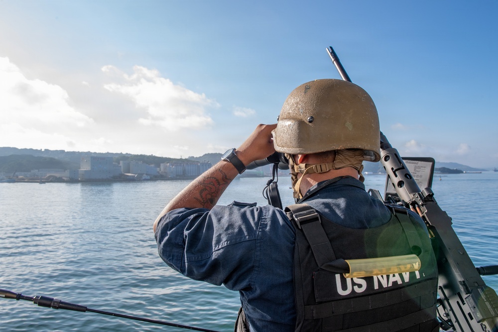 A US Navy sailor is standing on a vessel and looking through binoculars toward a coastal installation or port area. The sailor is wearing full tactical gear including a helmet, body armor vest marked 'US NAVY,' and is carrying a rifle on their back. The scene appears to be a harbor or bay approach, likely during a security or watch-standing operation.