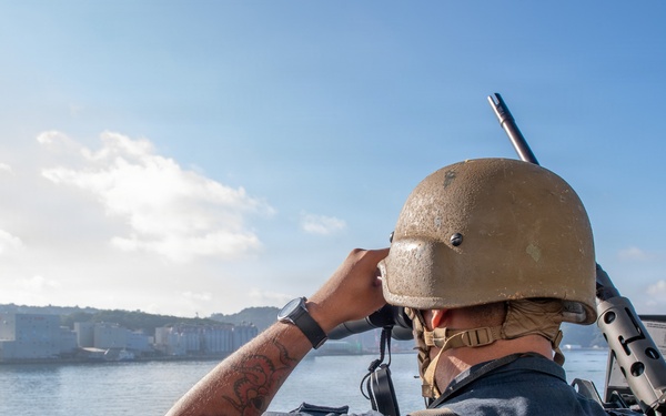 A US Navy sailor is standing on a vessel and looking through binoculars toward a coastal installation or port area. The sailor is wearing full tactical gear including a helmet, body armor vest marked 'US NAVY,' and is carrying a rifle on their back. The scene appears to be a harbor or bay approach, likely during a security or watch-standing operation.