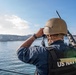 A US Navy sailor is standing on a vessel and looking through binoculars toward a coastal installation or port area. The sailor is wearing full tactical gear including a helmet, body armor vest marked 'US NAVY,' and is carrying a rifle on their back. The scene appears to be a harbor or bay approach, likely during a security or watch-standing operation.
