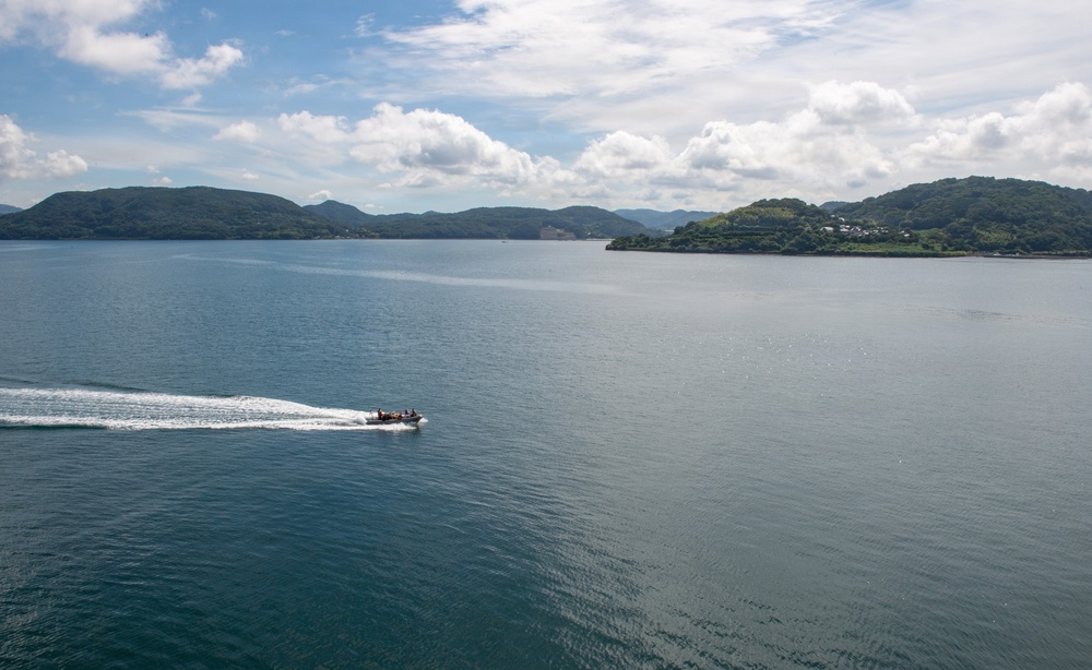 An aerial view of a calm bay or inlet surrounded by green, forested hills and islands. A single motorboat is cutting through the teal-blue water, leaving a white wake trail behind it. The scene appears to be in a temperate coastal region, possibly East Asia, with scattered buildings visible on the right shoreline.