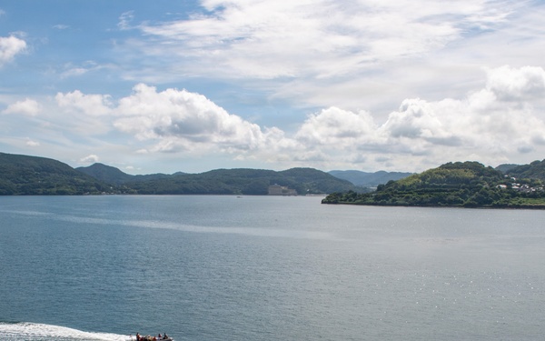 An aerial view of a calm bay or inlet surrounded by green, forested hills and islands. A single motorboat is cutting through the teal-blue water, leaving a white wake trail behind it. The scene appears to be in a temperate coastal region, possibly East Asia, with scattered buildings visible on the right shoreline.