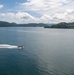 An aerial view of a calm bay or inlet surrounded by green, forested hills and islands. A single motorboat is cutting through the teal-blue water, leaving a white wake trail behind it. The scene appears to be in a temperate coastal region, possibly East Asia, with scattered buildings visible on the right shoreline.