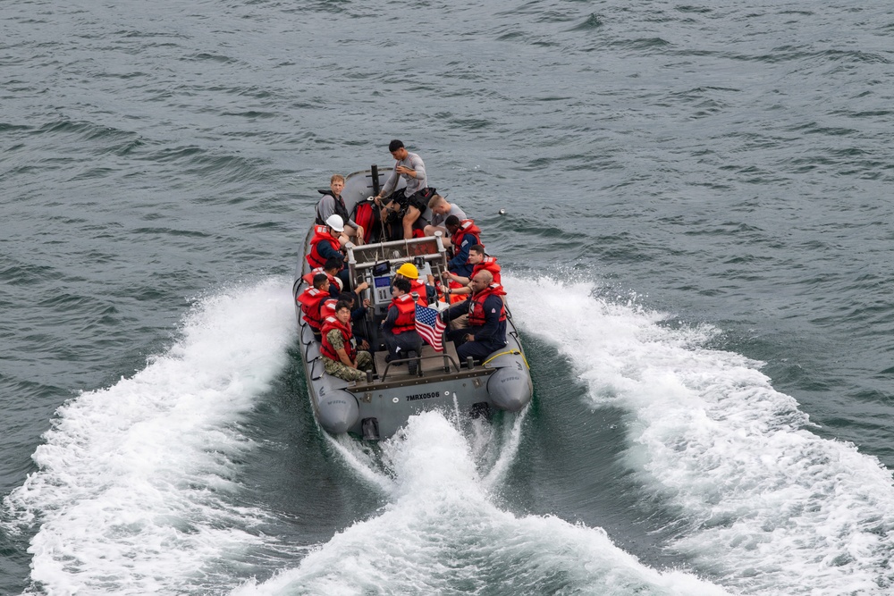 A rigid inflatable boat (RIB) is underway on open water, carrying approximately 10-12 personnel, several of whom are wearing red life vests. The boat displays a small American flag and has the hull number '7MRX0508' visible on its bow. The vessel appears to be a military or coast guard craft based on the personnel attire and equipment visible.