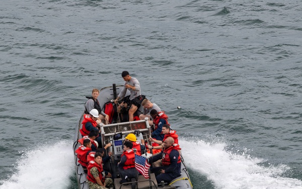 A rigid inflatable boat (RIB) is underway on open water, carrying approximately 10-12 personnel, several of whom are wearing red life vests. The boat displays a small American flag and has the hull number '7MRX0508' visible on its bow. The vessel appears to be a military or coast guard craft based on the personnel attire and equipment visible.