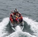 A rigid inflatable boat (RIB) is underway on open water, carrying approximately 10-12 personnel, several of whom are wearing red life vests. The boat displays a small American flag and has the hull number '7MRX0508' visible on its bow. The vessel appears to be a military or coast guard craft based on the personnel attire and equipment visible.