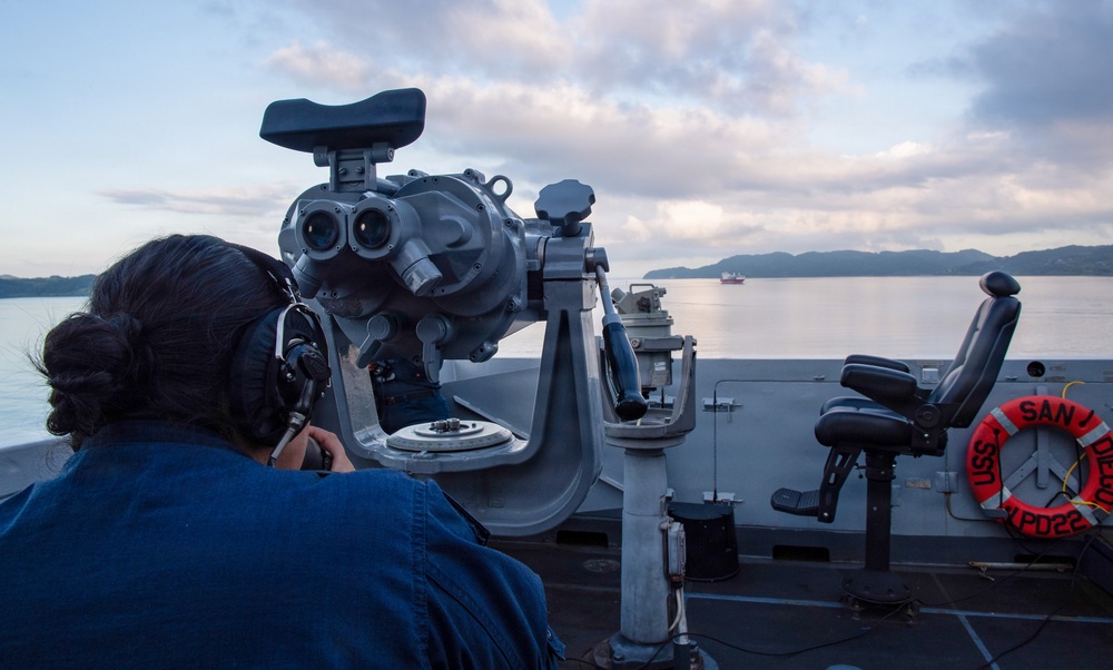 A naval personnel member is using large mounted binoculars (Big Eyes) on the bridge wing of a U.S. Navy ship, identified by the life ring as USS San Diego (LPD-22). The scene is set at dawn or dusk over calm coastal waters with hills visible in the background and a vessel in the distance. The sailor is wearing communications headgear while conducting a visual watch.