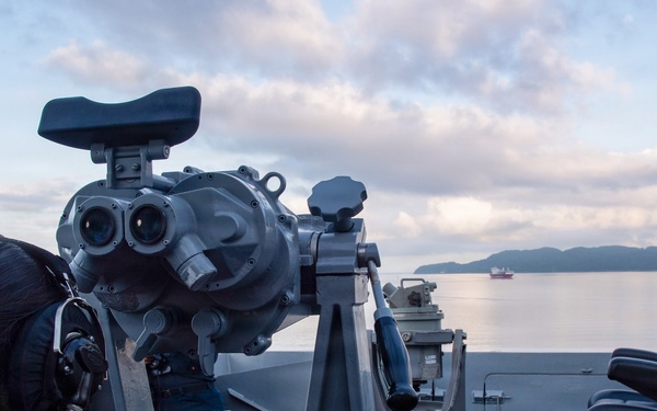 A naval personnel member is using large mounted binoculars (Big Eyes) on the bridge wing of a U.S. Navy ship, identified by the life ring as USS San Diego (LPD-22). The scene is set at dawn or dusk over calm coastal waters with hills visible in the background and a vessel in the distance. The sailor is wearing communications headgear while conducting a visual watch.