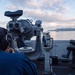 A naval personnel member is using large mounted binoculars (Big Eyes) on the bridge wing of a U.S. Navy ship, identified by the life ring as USS San Diego (LPD-22). The scene is set at dawn or dusk over calm coastal waters with hills visible in the background and a vessel in the distance. The sailor is wearing communications headgear while conducting a visual watch.